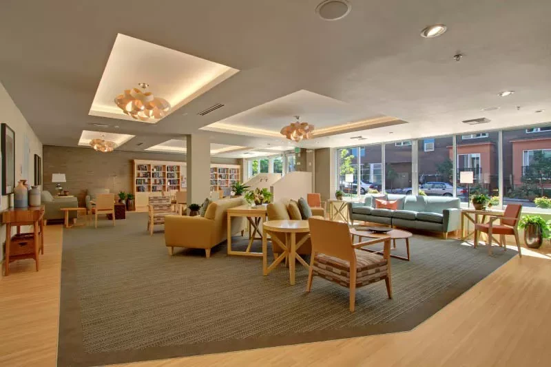 Modern community lounge with warm wood furnishings, mid-century style chairs arranged in conversational groupings, bookshelves along walls, large windows overlooking street, recessed ceiling lights with decorative fixtures, neutral carpet and hardwood flooring, and contemporary design throughout.