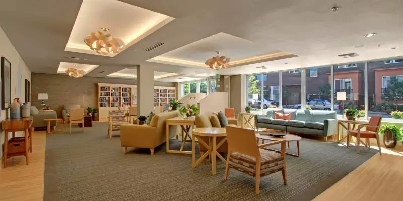 Modern community lounge with warm wood furnishings, mid-century style chairs arranged in conversational groupings, bookshelves along walls, large windows overlooking street, recessed ceiling lights with decorative fixtures, neutral carpet and hardwood flooring, and contemporary design throughout.