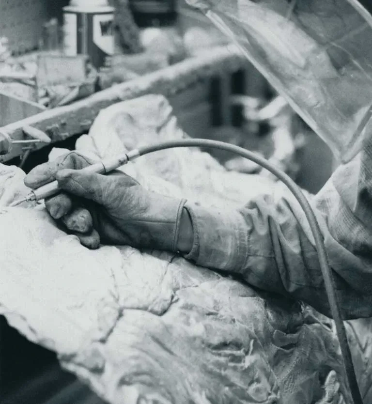 Black and white photograph showing gloved hands holding a curved pipe or hose over a textured surface, with blurred industrial or workshop equipment visible in the background.