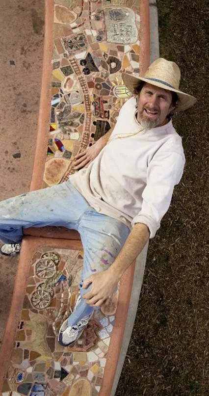 A man wearing a straw hat and white shirt lies on a colorful mosaic tile couch, smiling at the camera. The mosaic features intricate patterns with various colored tiles in geometric designs.