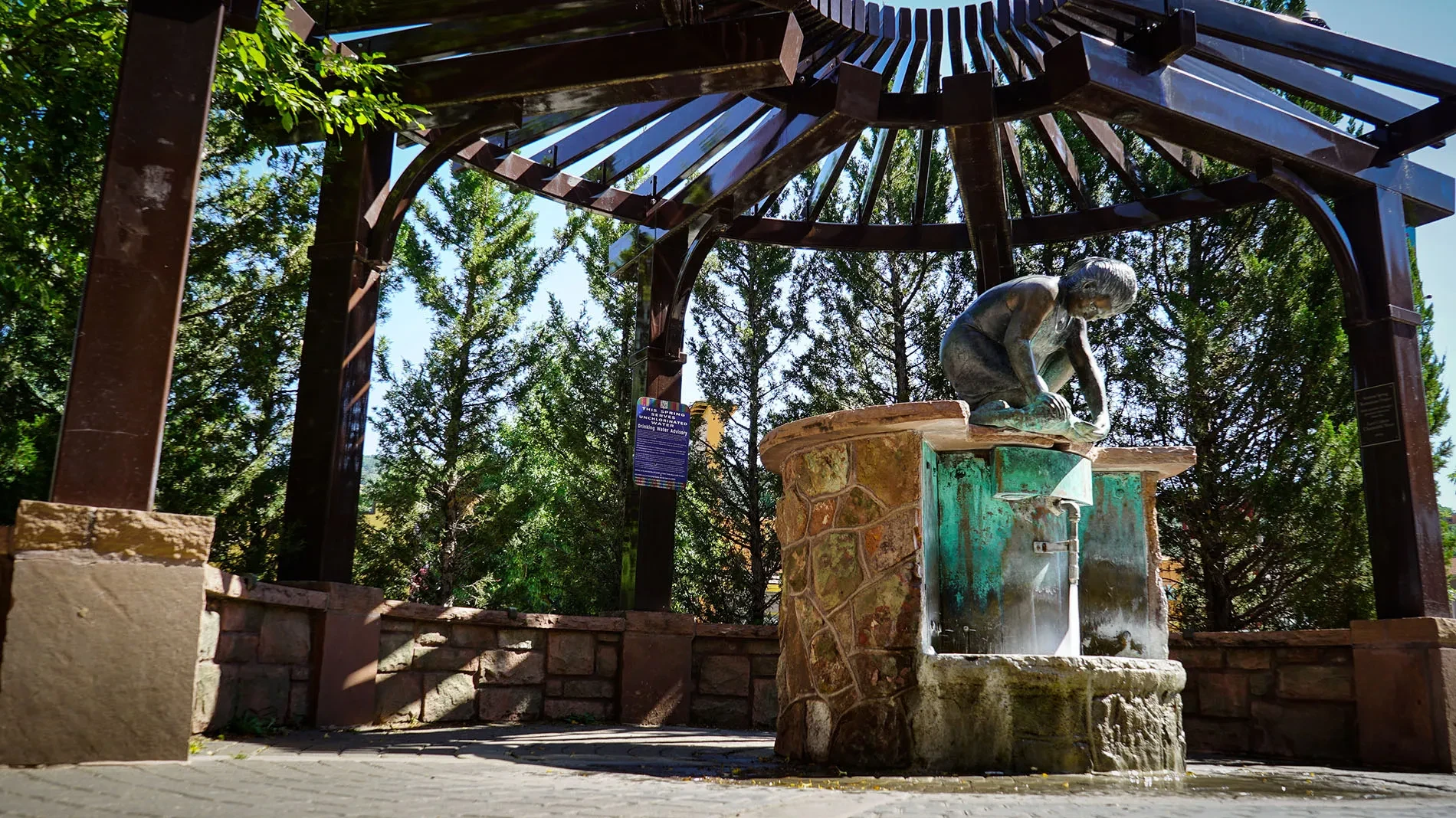 A bronze statue of a person leaning over a decorative stone basin sits beneath a dark wooden pergola structure. The figure appears to be reading or examining something in the basin. Tall green trees surround the area, and an informational plaque is visible on the left side. The setting is a shaded outdoor pavilion with clear skies visible above.