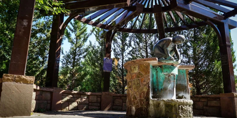 A bronze statue of a person leaning over a decorative stone basin sits beneath a dark wooden pergola structure. The figure appears to be reading or examining something in the basin. Tall green trees surround the area, and an informational plaque is visible on the left side. The setting is a shaded outdoor pavilion with clear skies visible above.